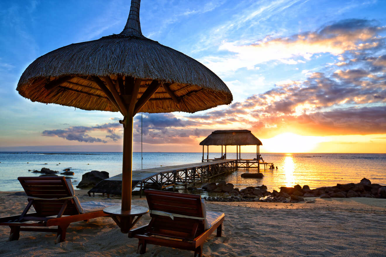 Sunset at a tranquil beach with a thatched umbrella and loungers in the foreground, leading to a wooden pier and gazebo over the water, under a sky aglow with the setting su